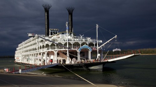 A large steamboat docked by the shore under a dark, cloudy sky, with two tall smokestacks and a red ramp.
