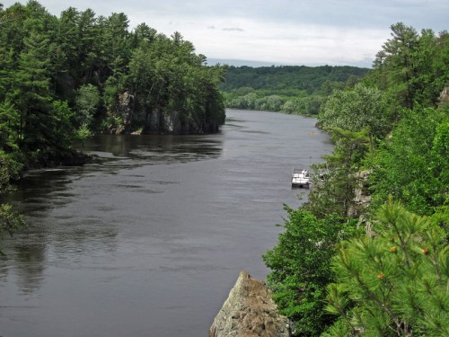 A serene river flows between lush green cliffs, with a small boat anchored nearby under a cloudy sky.