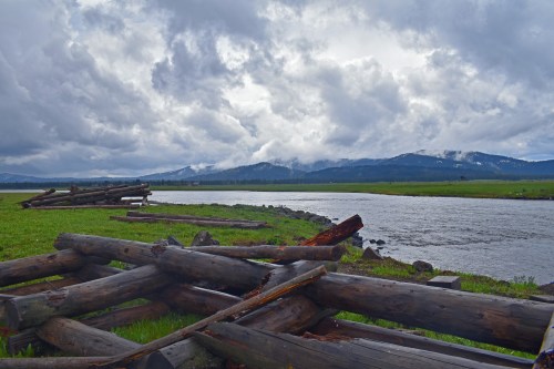 A serene river scene with logs in the foreground, mountains in the background, and cloudy skies above.