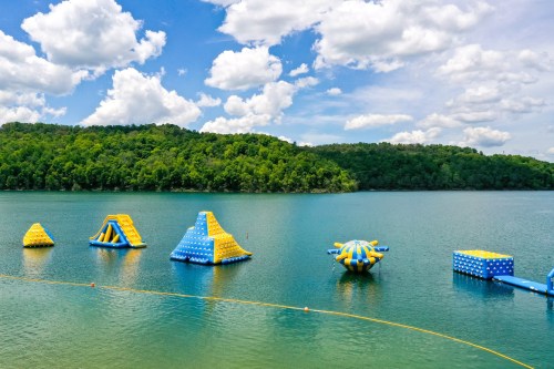 Colorful inflatable water obstacles on a lake, surrounded by green hills and a blue sky with fluffy clouds.