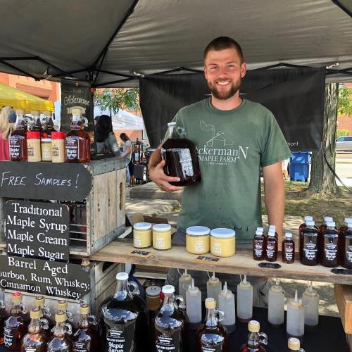 A smiling vendor holds a large jug of maple syrup at a market, surrounded by various maple products and a sign for free samples.