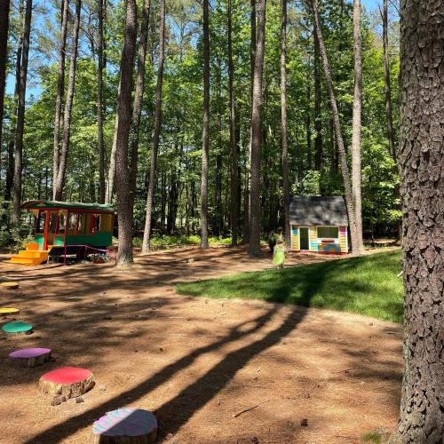 A colorful play area in a forest with a small house and a train-themed play structure among tall trees.