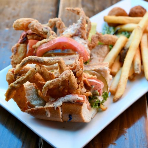 A sandwich with fried soft-shell crab, lettuce, and tomato, served with fries and a side of hush puppies.