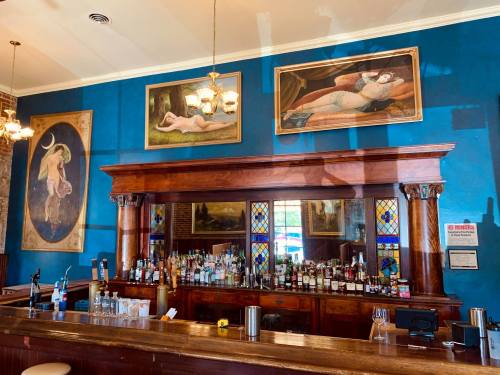 A vintage bar with a wooden counter, colorful stained glass, and framed art on a blue wall.