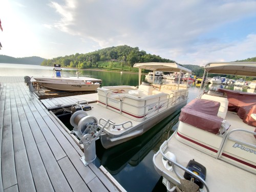 Two boats docked at a marina, surrounded by green hills and calm water under a partly cloudy sky.