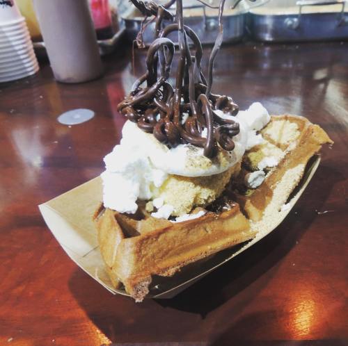 A waffle topped with whipped cream, cookie crumbles, and drizzled chocolate, served in a paper boat on a wooden table.