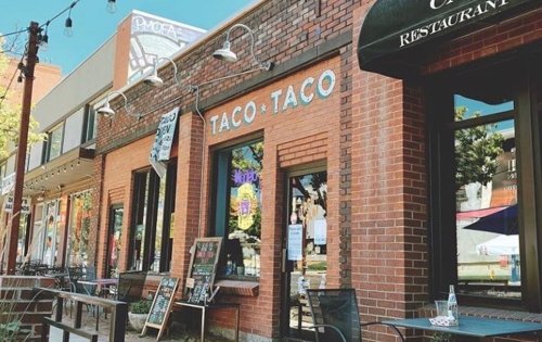 A brick building with "Taco Taco" sign, outdoor seating, and nearby shops on a sunny day.