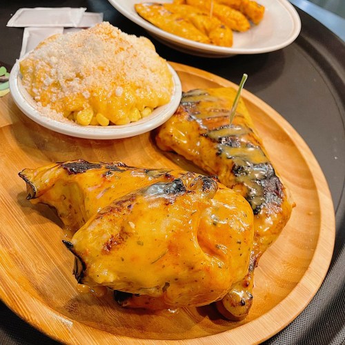 Grilled chicken pieces glazed with sauce, served with a side of creamy corn and fried plantains on a wooden platter.