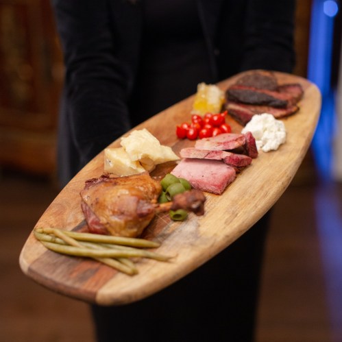 A wooden platter holds various meats, pickles, and garnishes, presented by a person in a black outfit.