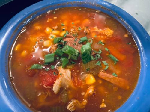 A blue bowl filled with hearty soup featuring chicken, corn, tomatoes, and green onions, garnished with fresh herbs.