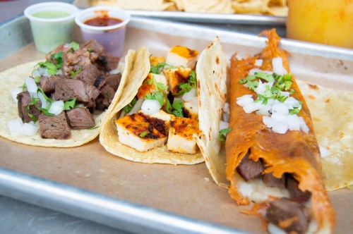 Three tacos on a tray: one with beef, one with grilled chicken, and a third with beef and red sauce, garnished with onions and cilantro.
