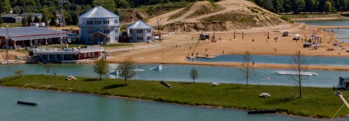 A scenic view of a lake with sandy beach, buildings, and people enjoying outdoor activities in a recreational area.
