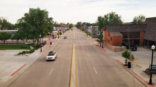 A wide, empty street lined with trees and buildings under a cloudy sky, with a few cars parked along the sides.