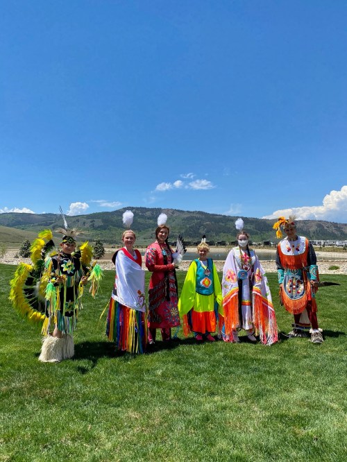 A group of six individuals in traditional attire stands on green grass with mountains and blue sky in the background.