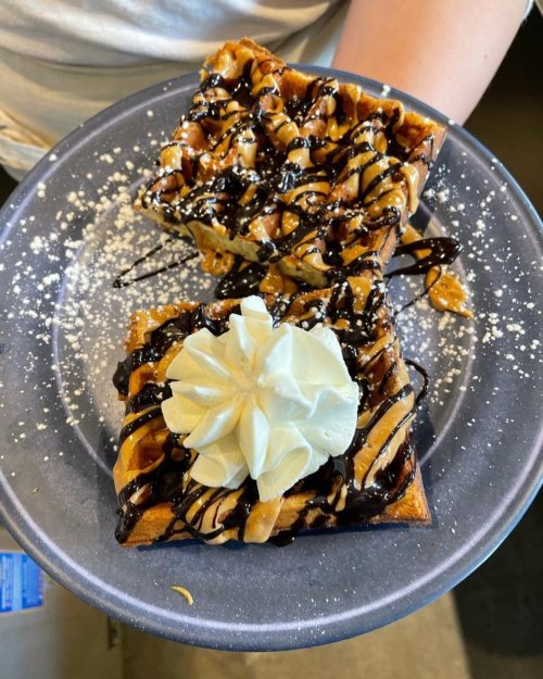 A plate of two waffles topped with chocolate drizzle, caramel, nuts, and a dollop of whipped cream, dusted with powdered sugar.