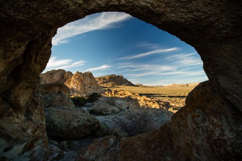 View through a rock formation, showcasing a desert landscape with mountains and a blue sky.