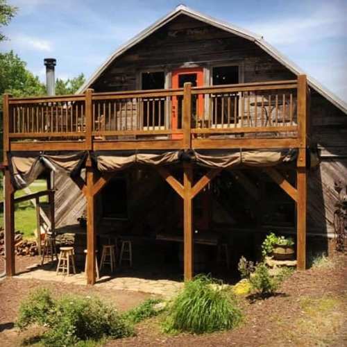A rustic wooden cabin with a large deck, surrounded by greenery and a stone pathway.