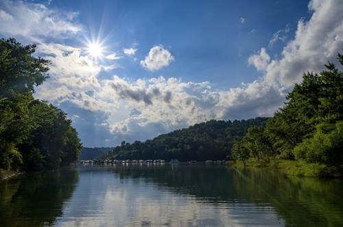 A serene lake scene with boats, surrounded by lush greenery and a bright sun shining through scattered clouds.