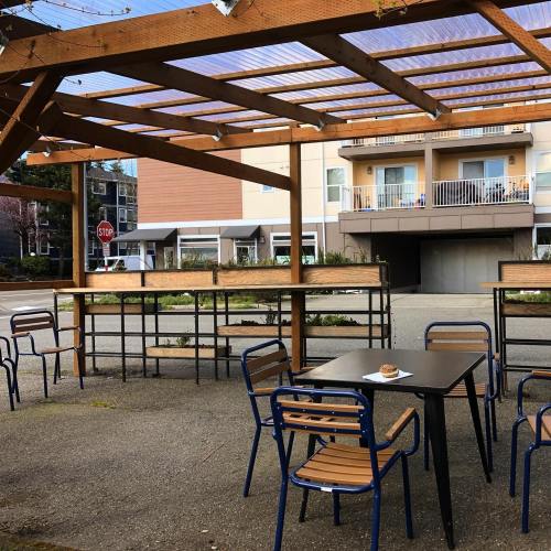 Outdoor seating area with wooden pergola, tables, and chairs, near a building with balconies and a stop sign.