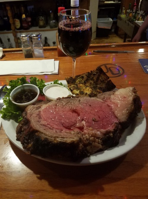 A plate of prime rib with sides of sauce and a glass of red wine on a wooden bar counter.