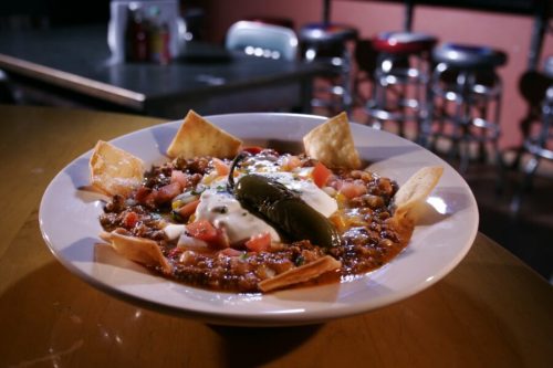 A bowl of chili topped with sour cream, jalapeño, tomatoes, and tortilla chips, served on a wooden table.