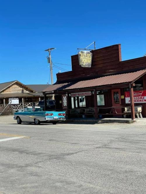 A vintage blue car parked in front of a rustic pizza restaurant under a clear blue sky.
