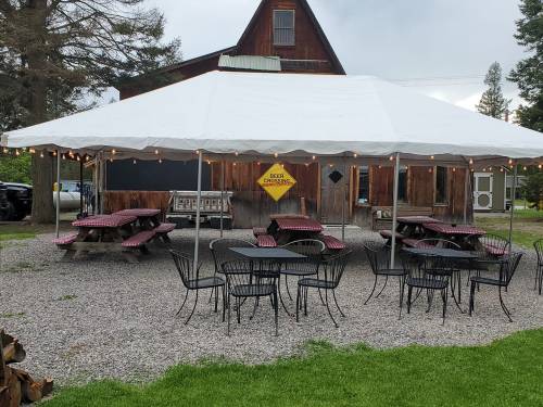 Outdoor seating area with picnic tables and black chairs under a large tent near a rustic wooden building.