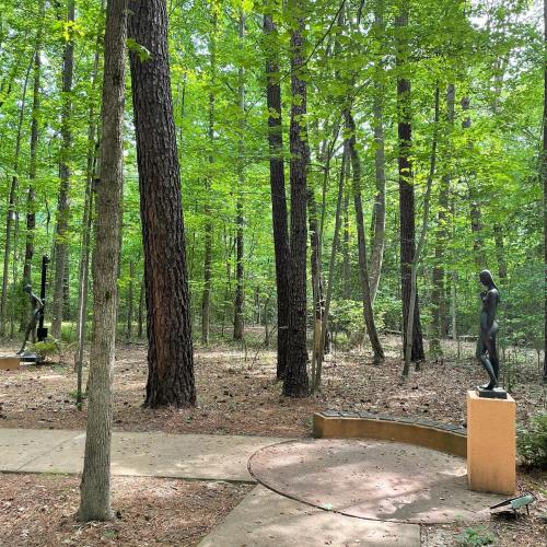 Two sculptures stand on pedestals in a lush green forest, surrounded by tall trees and dappled sunlight.