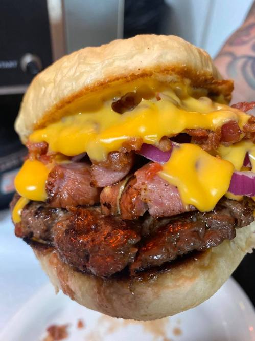 A close-up of a loaded burger with layers of cheese, bacon, and meat, held in a hand against a blurred background.