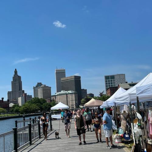 A bustling waterfront market with people walking, tents set up, and city buildings in the background under a clear blue sky.