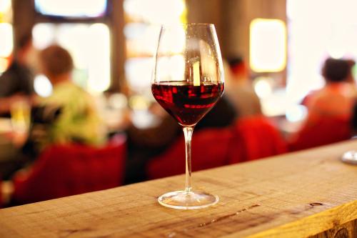 A glass of red wine sits on a wooden bar, with blurred figures in the background.