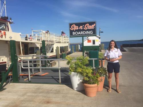 A woman stands by a boarding sign for "Sip n' Salt" near a ferry dock on a sunny day.