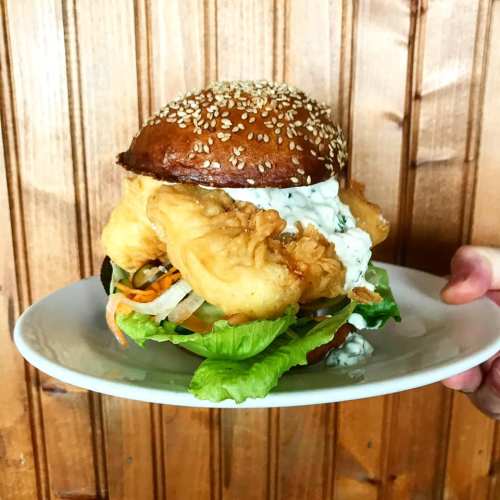 A hand holding a plate with a large, crispy fish burger topped with lettuce, vegetables, and a creamy sauce on a sesame bun.