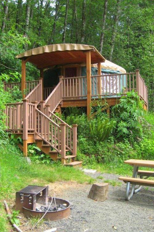 A cozy yurt on a wooden deck surrounded by lush greenery, with a fire pit and picnic table nearby.