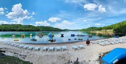 A scenic lake view with inflatable water toys, sandy beach, and lounge chairs under a blue sky with fluffy clouds.