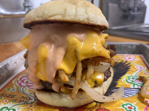 A towering burger with melted cheese, fries, grilled onions, and a creamy sauce, served on a colorful tray.