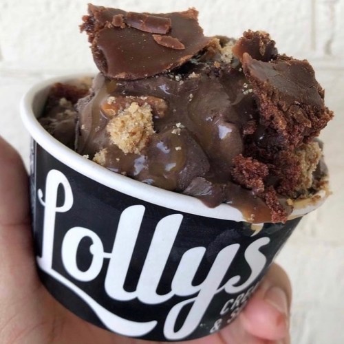A hand holding a cup of chocolate ice cream topped with chunks of brownie and cookie pieces.