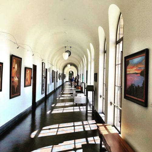 A long, bright hallway with arched windows, featuring artwork on the walls and people walking in the distance.