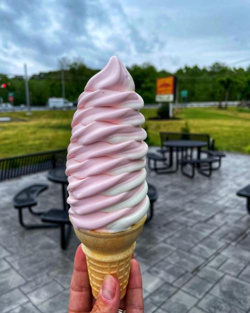 A hand holding a large swirl of pink and white soft serve ice cream in a cone, with outdoor seating in the background.