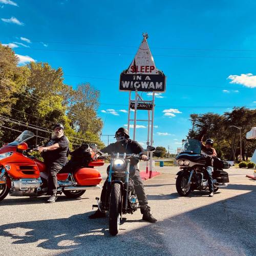 Three motorcyclists pose in front of a vintage "Sleep in a Wigwam" sign on a sunny day.