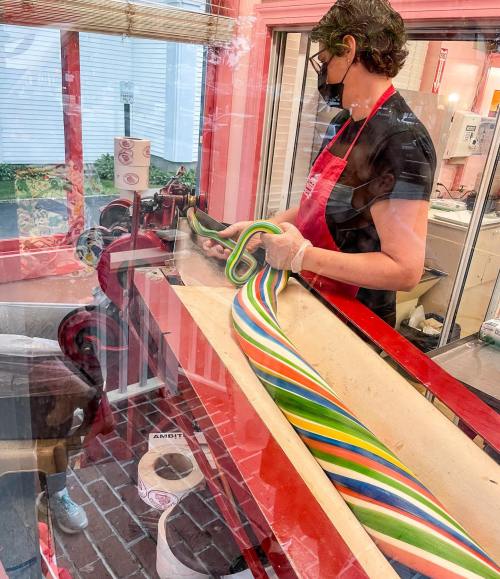 A person in an apron uses a machine to shape colorful candy in a shop, with vibrant stripes of candy visible.
