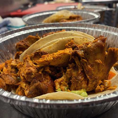 Close-up of tacos filled with seasoned meat, served in aluminum trays with toppings like guacamole and salsa.