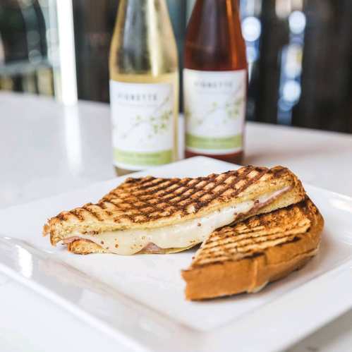 A grilled sandwich with melted cheese and ham, served on a white plate with two bottles of wine in the background.
