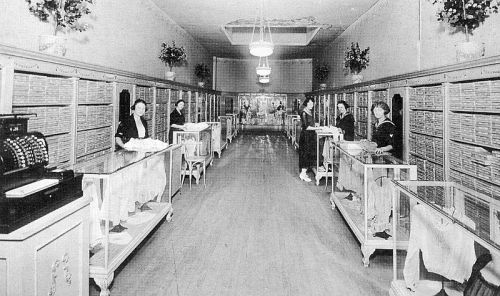 A vintage store interior with women working at display tables and a cash register, surrounded by shelves of merchandise.