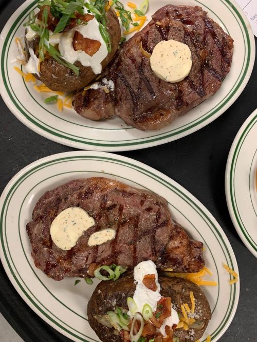 Two plates of grilled steak with herb butter, served with loaded baked potatoes topped with sour cream and green onions.
