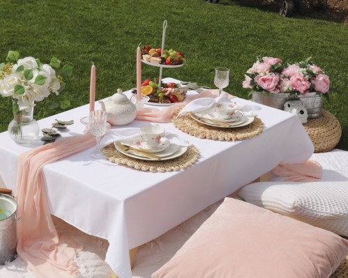 A beautifully set outdoor table with floral arrangements, elegant tableware, and a tiered stand of fruits and desserts.