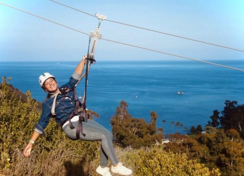 A person zip-lining over a scenic coastal view, smiling with the ocean and trees in the background.
