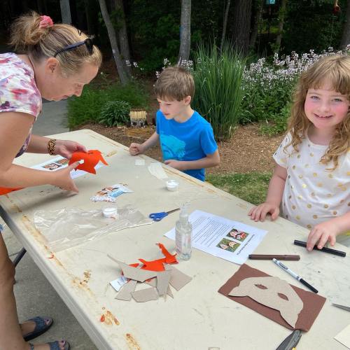 A woman and two children craft with paper and glue at an outdoor table surrounded by greenery.