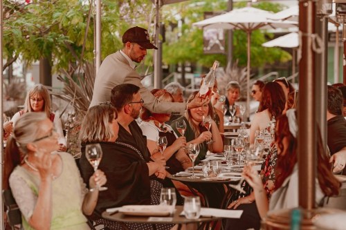 A lively outdoor restaurant scene with diners enjoying drinks, a waiter serving wine, and greenery in the background.