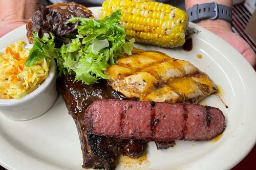 A plate with grilled chicken, sausage, ribs, corn on the cob, and a side of coleslaw and mixed greens.
